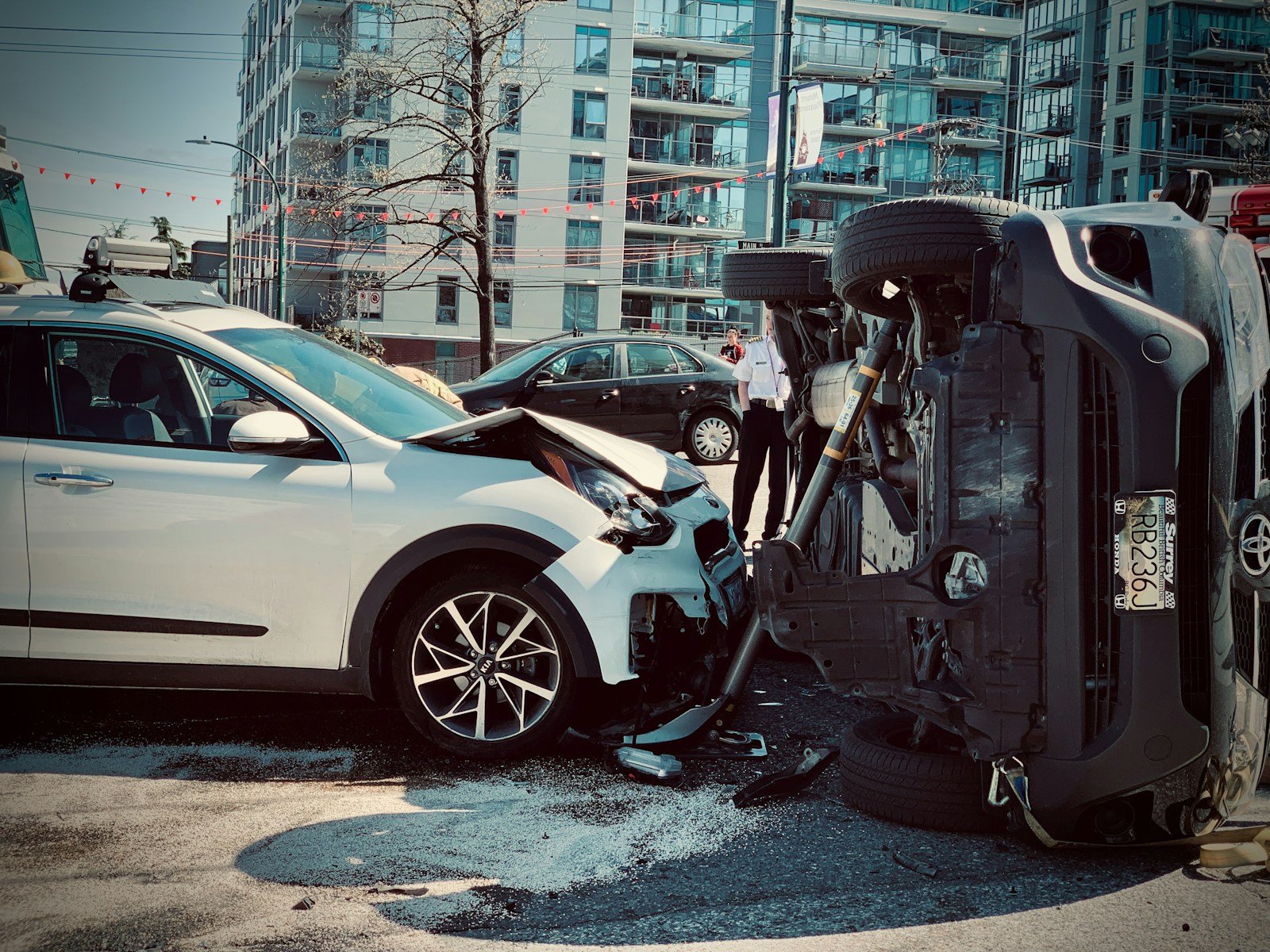 A damaged vehicle at an accident scene with a second overturned car, bystanders, and visible license plates in the background